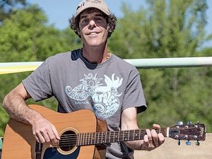 A person wearing a cap and a t-shirt with a graphic design is playing an acoustic guitar outside. The background includes green trees and a railing, suggesting an outdoor setting on a sunny day. The person is smiling and appears to be enjoying the moment. Fearrington Village