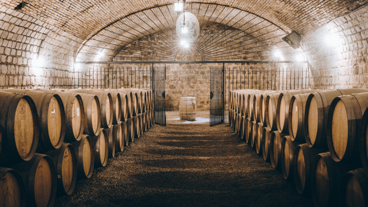 A dimly lit wine cellar with arched brick ceilings, stone walls, and hanging light fixtures. Wooden barrels are neatly arranged in rows on both sides, leading to a single barrel at the far end, creating a sense of depth and symmetry. Gravel covers the floor. Fearrington Village