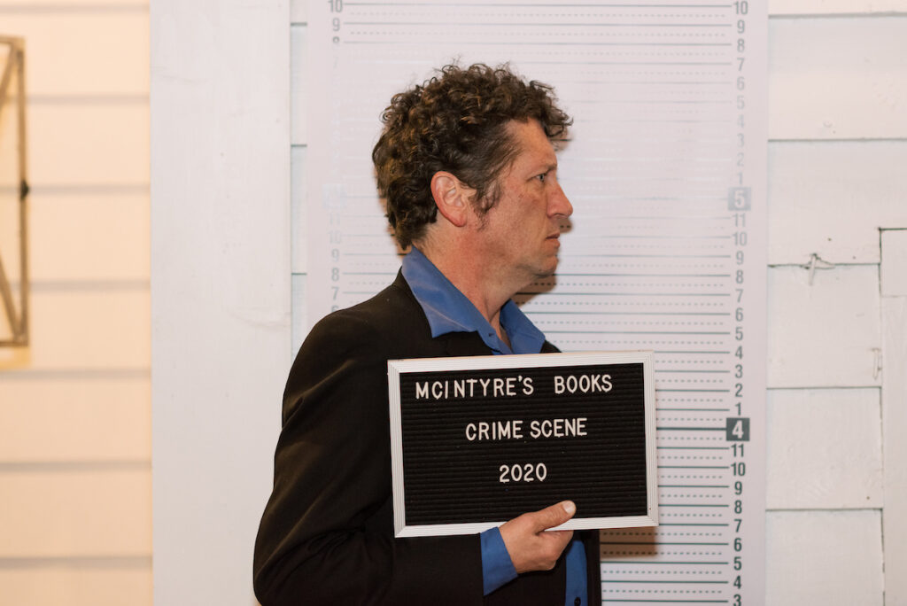 A man with curly hair and a serious expression is holding a black sign that reads "McIntyre's Books Crime Scene 2020." He stands in front of a height measurement chart, appearing as if in a staged mugshot photo. He is wearing a black jacket and blue shirt. Fearrington Village