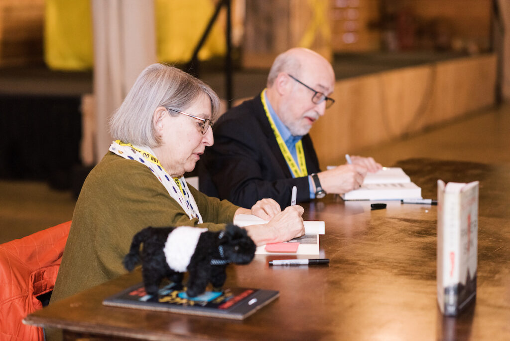 Two elderly people, sitting at a table, are visible signing autographs or books. Both are wearing glasses and lanyards, with a small stuffed animal on the table. The background is blurred, suggesting an indoor event setting. Fearrington Village