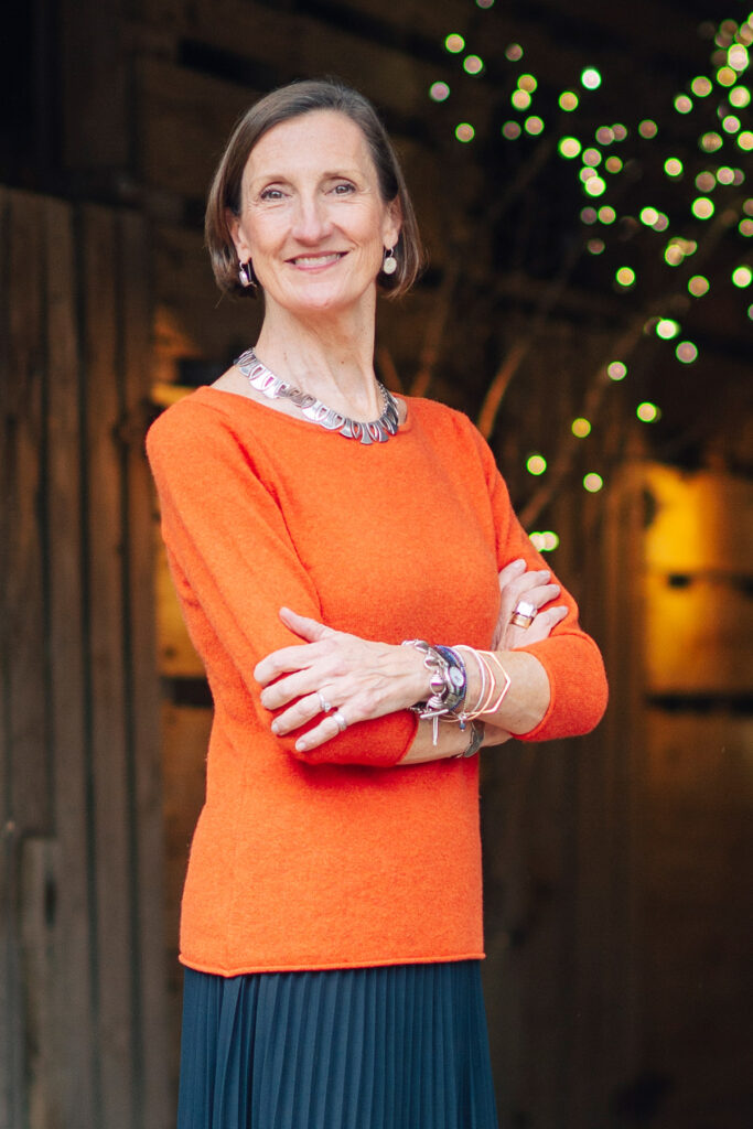 A woman stands confidently with crossed arms, smiling at the camera. She is wearing an orange sweater, a statement necklace, and bracelets. She has chin-length hair tucked behind her ears. The background features wooden panels and a warm string of lights. Fearrington Village