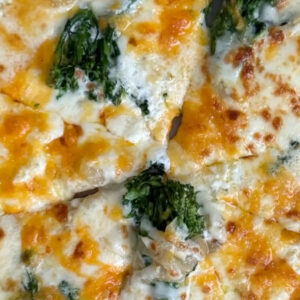 A close-up of a sliced pizza topped with melted cheese and green broccoli florets. The crust appears thin and crispy, and the cheese is golden brown with some slightly browned areas. Fearrington Village