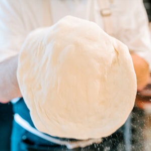 A person wearing a white shirt is tossing a large, stretchy pizza dough in the air. The focus of the image is on the dough, which appears soft and slightly translucent. The person's face and background are out of focus. Fearrington Village