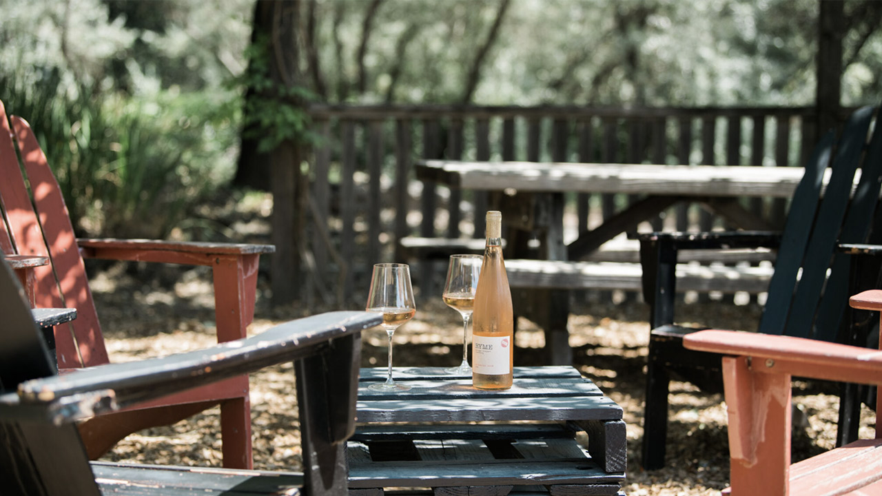 A tranquil outdoor setting featuring a wooden table with two wine glasses and a bottle of white wine. Surrounding the table are colorful Adirondack chairs, and a wooden fence with lush greenery in the background. Fearrington Village