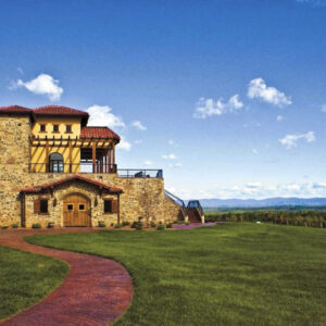 A large, rustic-style house with stone walls, red tile roofs, and wooden accents sits on a vast, green lawn. A winding brick pathway leads to the entrance. The background features expansive hilly terrain under a clear blue sky with scattered clouds. Fearrington Village