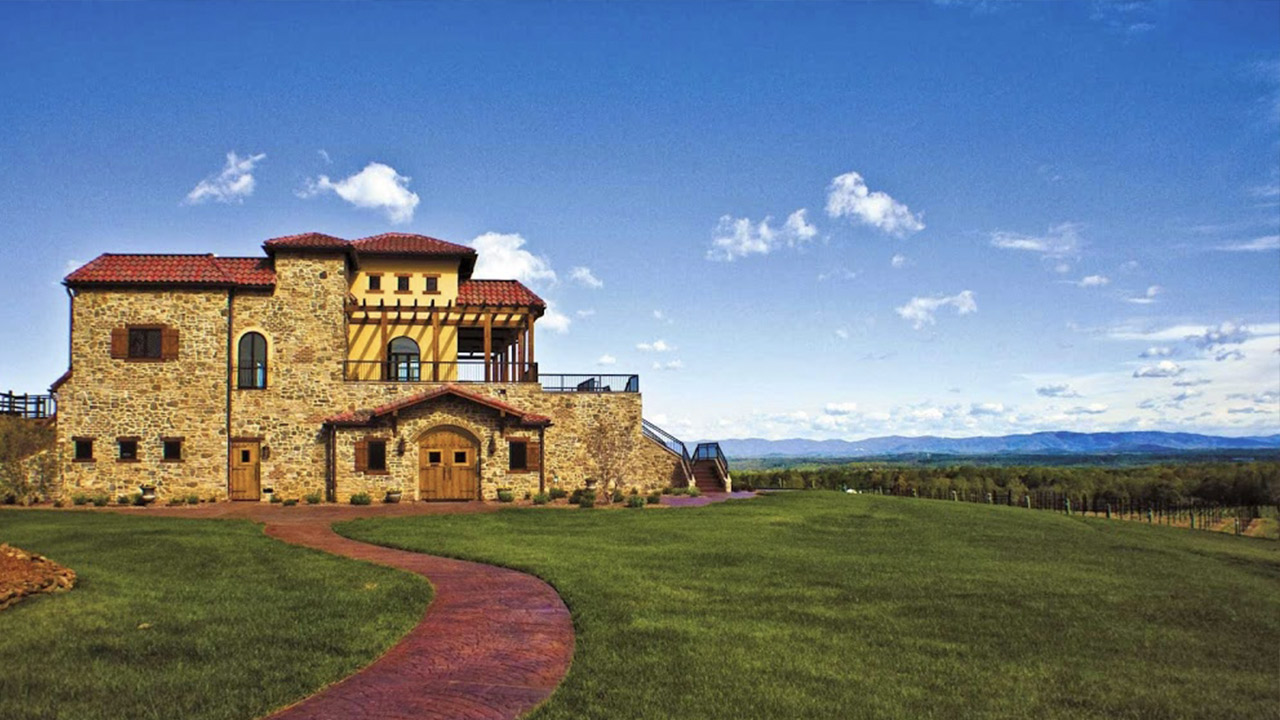 A large, rustic-style house with stone walls, red tile roofs, and wooden accents sits on a vast, green lawn. A winding brick pathway leads to the entrance. The background features expansive hilly terrain under a clear blue sky with scattered clouds. Fearrington Village