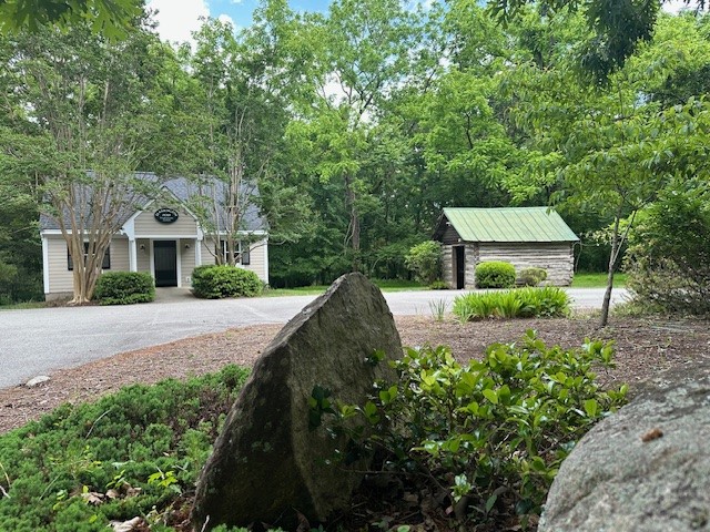 You've Got Mail! 1 A quaint building with a porch and a wooden shed with a green metal roof are surrounded by lush green trees and foliage. In the foreground, large stones and bushes are part of a landscaped garden. The sky is partly cloudy. Fearrington Village