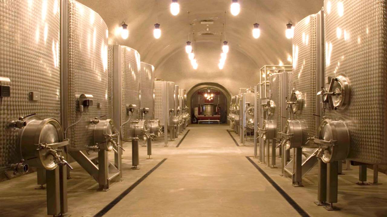 A large, well-lit wine cellar with a high arched ceiling. Stainless steel fermentation tanks line both sides of a central walkway, leading to a warmly glowing room in the distance. The tanks are cylindrical, with various valves and gauges attached. Fearrington Village