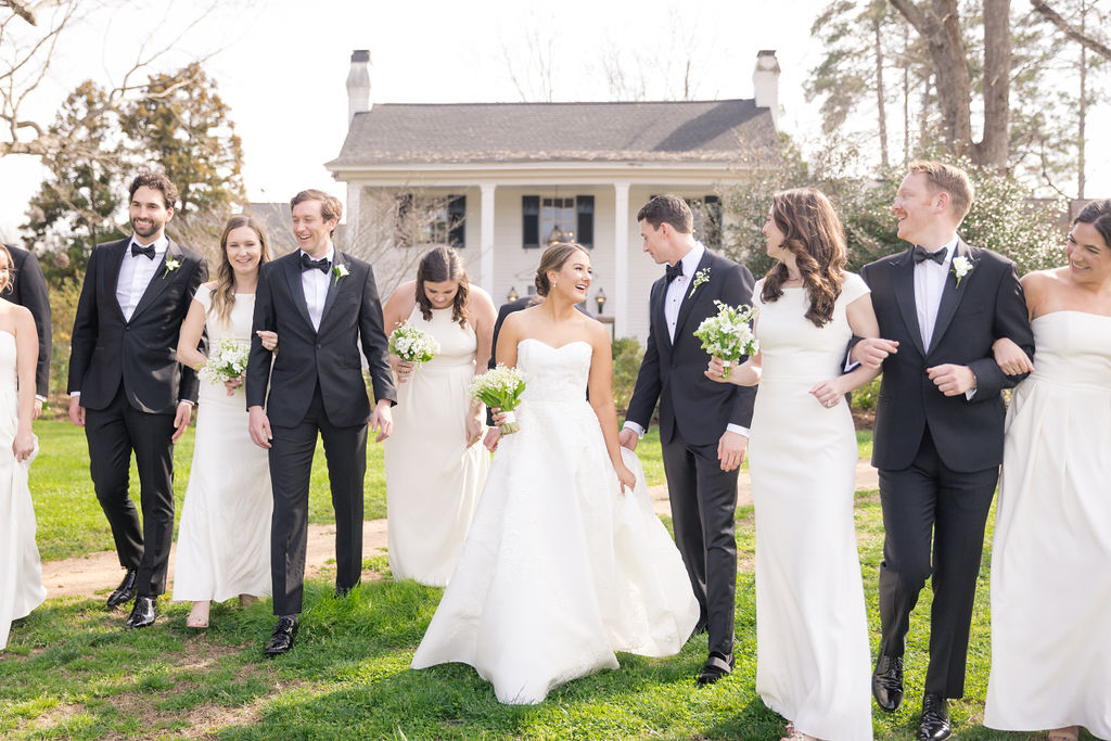 A bride and groom walk hand in hand, surrounded by six other wedding party members. The group, all dressed in elegant formal attire with bouquets, walks outdoors on a sunny day, with a charming, rustic house in the background. Everyone appears joyful and smiling. Fearrington Village