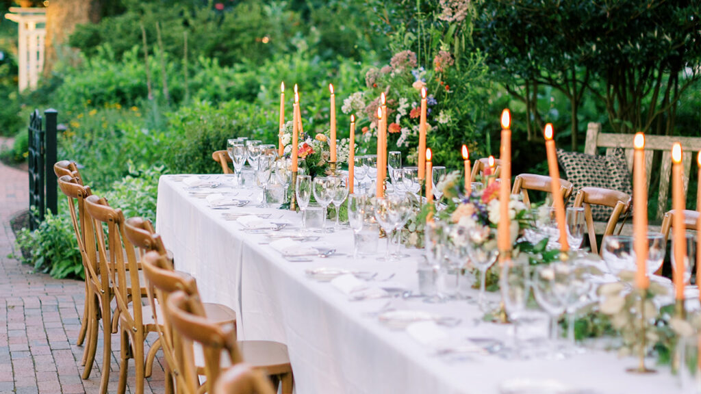 An elegantly set outdoor dining table with a white tablecloth is adorned with flower arrangements, tall lit candles, glassware, and silverware. Wooden chairs are neatly arranged along both sides of the table, and lush greenery forms a picturesque backdrop. Fearrington Village