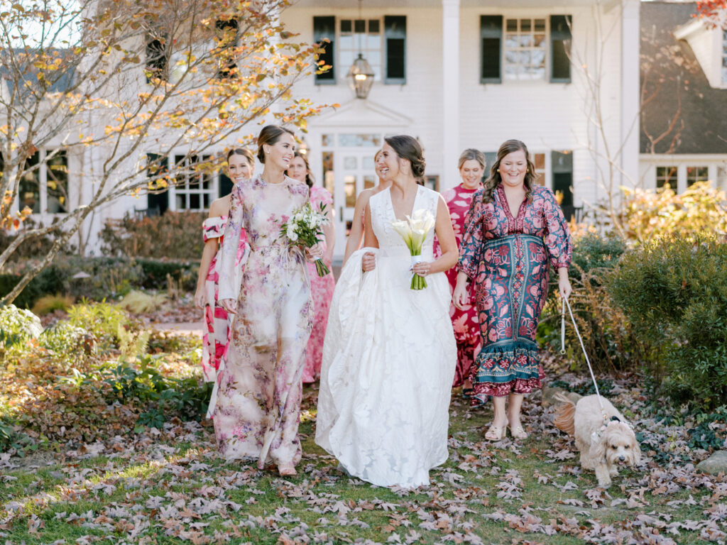 A bride in a white gown holding a bouquet walks outdoors with her bridal party. The bridesmaids wear colorful floral dresses, and one bridesmaid walks a small dog on a leash. They are all smiling and walking on a leaf-covered path with a house in the background. Fearrington Village