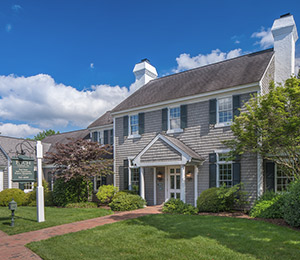 A two-story, colonial-style house with grey shingles, white trim, and green shutters, surrounded by well-maintained greenery and a brick pathway leading to the entrance. A white sign with a green face is located on the lawn near the sidewalk. The sky is clear and blue. Fearrington Village