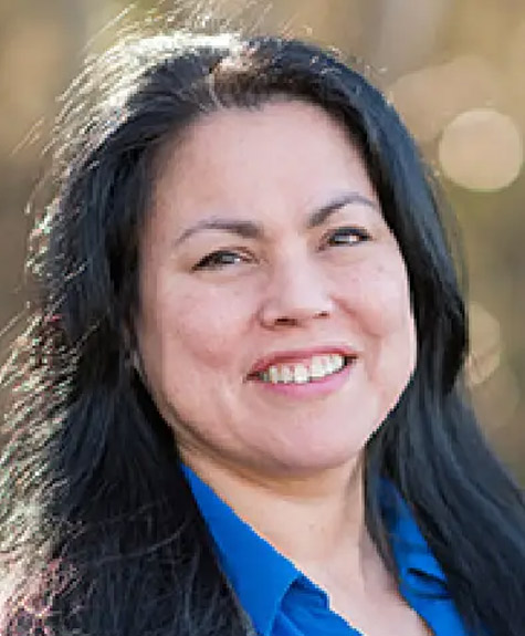 A person with long, dark hair and a friendly smile is seen in a close-up outdoor portrait. They are wearing a blue collared shirt, and the background is blurred with natural, warm tones, suggesting a sunny day. Fearrington Village