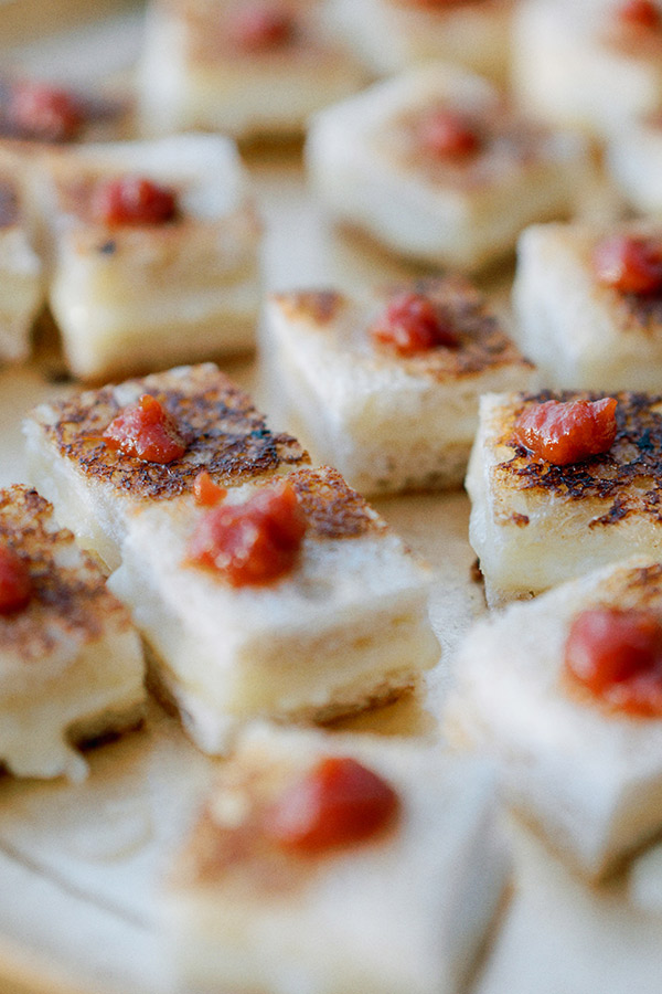 Close-up image of neatly arranged bite-sized tofu squares with a golden-brown seared surface, topped with small dollops of red sauce. The tofu pieces are placed close to each other on a flat surface. Fearrington Village
