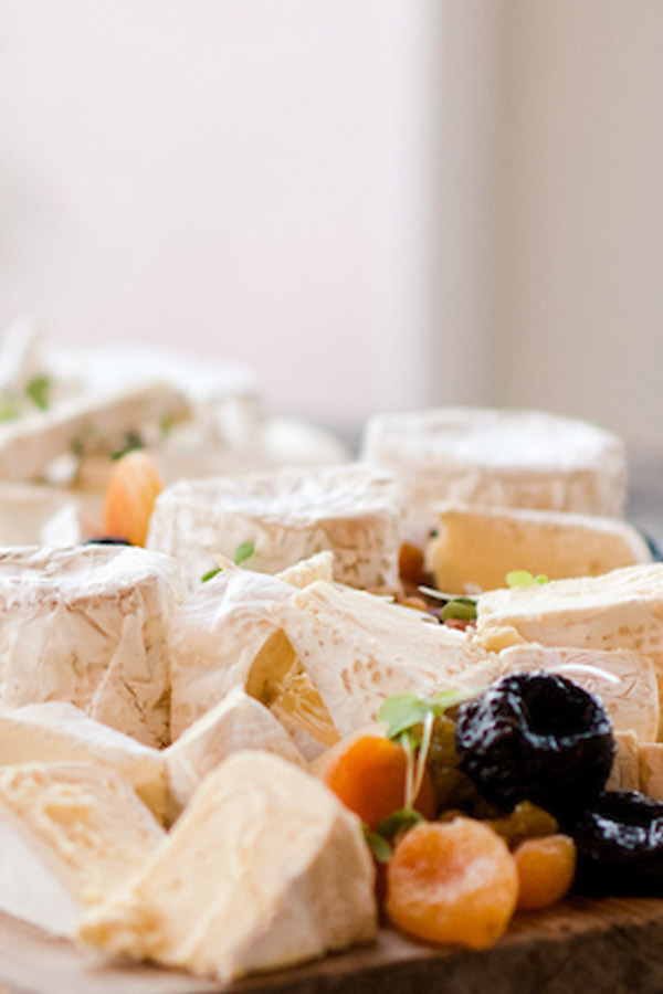 Close-up of a cheese platter featuring various types of soft cheeses with white rinds, accompanied by dried fruits such as apricots and prunes. The foods are arranged on a wooden board, creating an appetizing and gourmet presentation. Fearrington Village