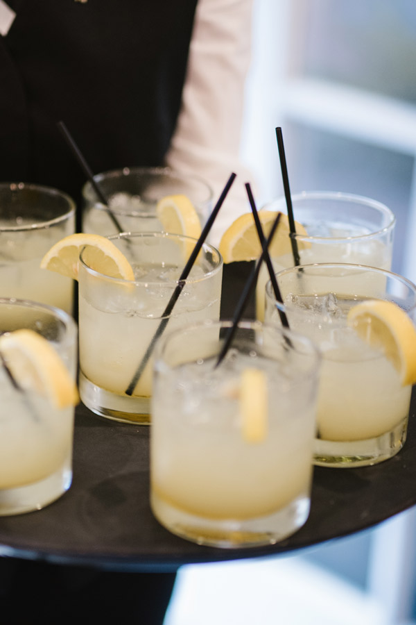 A server holds a tray filled with several glasses of a yellow beverage, each garnished with a slice of lemon and a black straw. The drinks appear to be on the rocks, with visible ice cubes in each glass. The server wears a white shirt and black vest. Fearrington Village