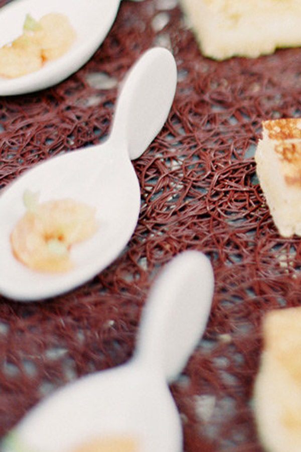 Close-up of a decorative table setting featuring small appetizers. The image shows white, spoon-shaped dishes holding bite-sized food, including shrimp, against a textured, burgundy placemat. Focus is on the front spoon, with additional blurred appetizers in the background. Fearrington Village