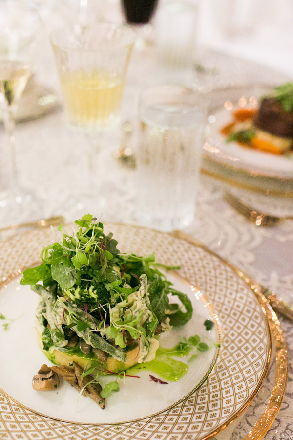 A gourmet dish featuring a fresh green salad with microgreens is served on an elegant, patterned plate. The table is set with crystal glasses filled with various beverages and intricate dinnerware, with another plated dish visible in the background. Fearrington Village