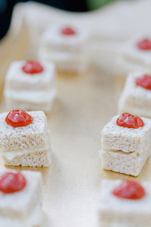 Close-up of small, square, layered finger sandwiches topped with a dollop of red sauce on a beige surface. The sandwiches appear to be made with white bread and a creamy white filling. Fearrington Village