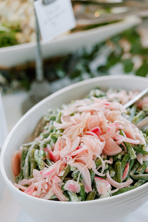 A white oval dish filled with green beans and topped with thinly sliced pink pickled onions, garnished and mixed with a creamy dressing. A spoon is placed in the dish for serving. In the background, another dish is slightly blurred. Fearrington Village