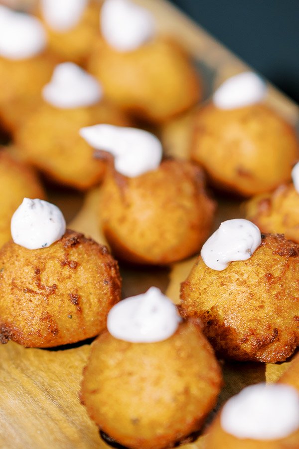 Close-up of multiple golden-brown hush puppies topped with small dollops of creamy white sauce, displayed on a wooden surface. The hush puppies are in focus with a blurry background, emphasizing their crisp texture and appealing presentation. Fearrington Village