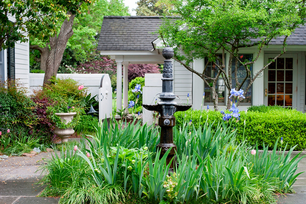 A charming garden scene featuring a vintage black water fountain surrounded by lush green plants and vibrant flowers. In the background, there is a quaint gray house with white trim, partially obscured by greenery and blooming trees. A walkway leads to the house. Fearrington Village