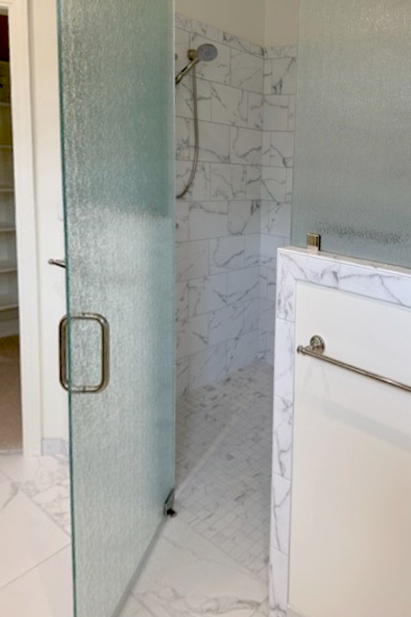 A bathroom with a marble-tiled shower area featuring a frosted glass door that is partially open. The showerhead is mounted on the marble wall. Outside the shower, there's a marble countertop with a towel rail attached to the side. Fearrington Village