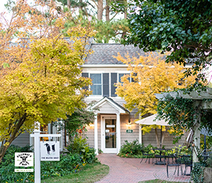 A charming building with beige siding is partially obscured by trees with yellow leaves. A brick path leads to the entrance, and there are outdoor tables with umbrellas to the right. A white sign with a dog graphic stands in the garden to the left of the entrance. Fearrington Village