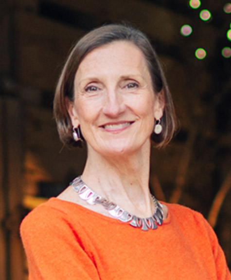 A smiling woman with short, brown hair is wearing an orange top, a silver necklace, and matching earrings. She is standing indoors with blurred background lights, suggesting a warm, cozy atmosphere. Fearrington Village