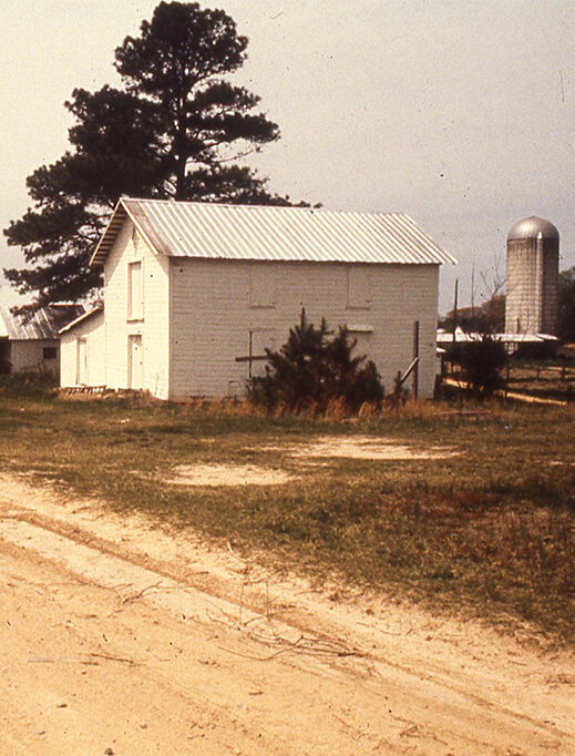 An old white barn with a metal roof stands in a rural area, surrounded by grass and dirt. A tall silo is visible in the background, along with a large tree next to the barn. The scene is bathed in warm sunlight, evoking a peaceful, rustic atmosphere. Fearrington Village