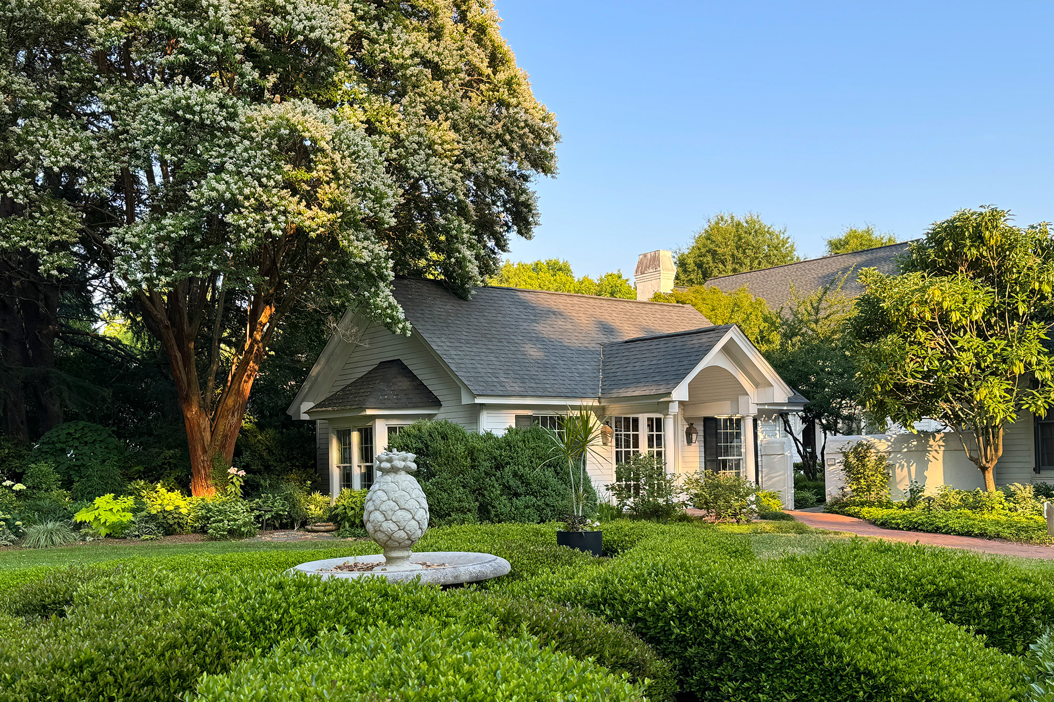 A charming house with a gray shingled roof and white trim sits amidst lush greenery. A pineapple-shaped fountain centerpiece stands in the well-manicured front garden, bordered by neatly trimmed hedges. Tall trees provide shade in the background under a clear blue sky. Fearrington Village