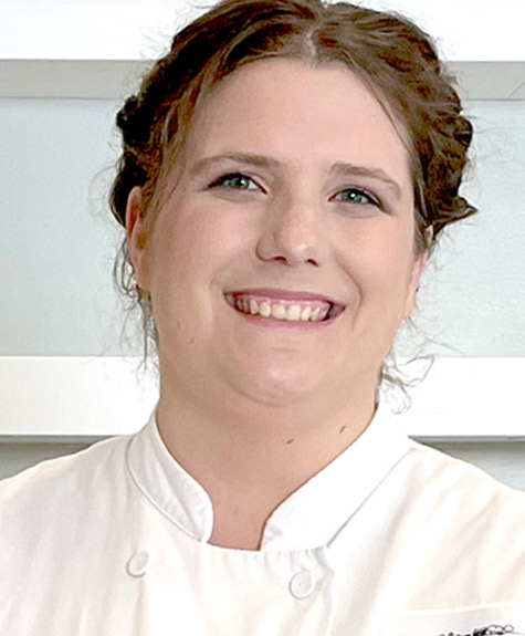 A smiling person with brown hair styled in a loose updo is wearing a white chef's coat. The background includes white shelves against a light-colored wall. Fearrington Village