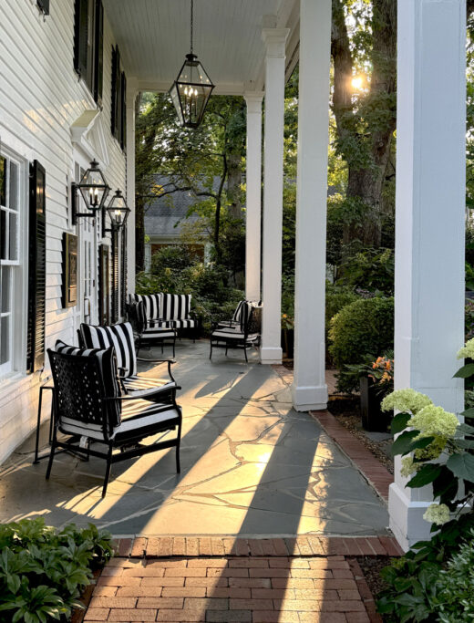 A sunlit porch with white columns and black-and-white striped cushions on black metal chairs. The stone floor is shaded by overhanging trees, and the evening sun casts long shadows. Lush greenery and white hydrangeas surround the area. Fearrington Village