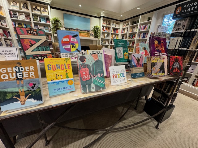 A display table in a bookstore showcases various books, including "Gender Queer," "The Guncle," "Heartstopper," "The Seven Husbands of Evelyn Hugo," and "Transmission." Shelves filled with more books are visible in the background. Fearrington Village