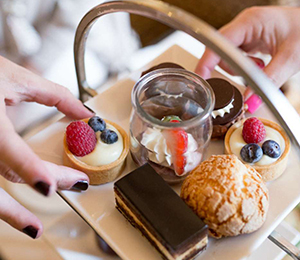 A close-up of a dessert tray with a variety of pastries and sweets. There are mini fruit tarts topped with raspberries and blueberries, a small glass jar with strawberry-topped cream, a chocolate tart, a cream puff, and a chocolate layered cake being held by two hands. Fearrington Village