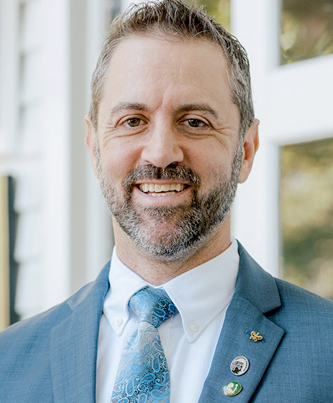 A man with short brown hair and a beard is smiling. He is wearing a blue suit, white shirt, and a blue paisley tie. He has several pins on his lapel. The background is blurred but shows a white structure with windows. Fearrington Village