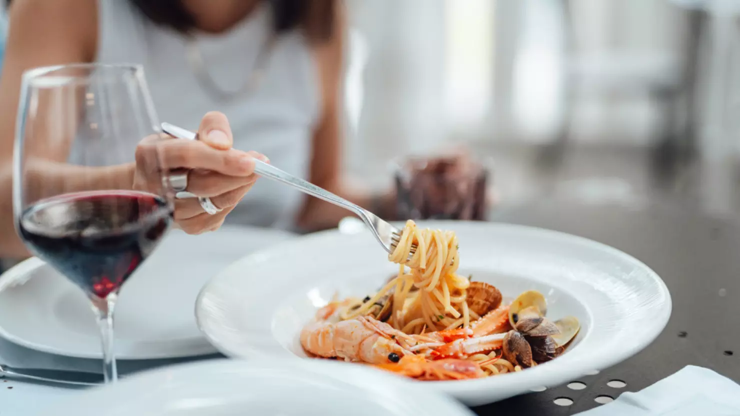 A person in a white top is twirling a forkful of pasta from a plate of seafood linguine. The dish features shrimp, clams, and other shellfish. A glass of red wine is on the table beside the plate. The scene is set in a well-lit dining area. Fearrington Village