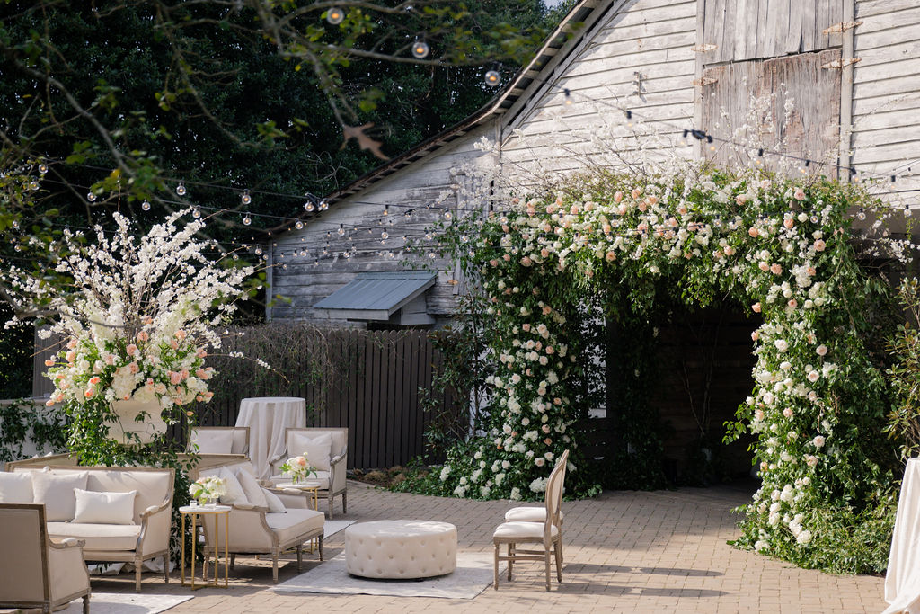 Rustic outdoor wedding venue with a wooden barn adorned with lush floral arrangements of white and pink roses. Elegant white seating is arranged on a brick patio, featuring couches, chairs, and a round ottoman. Hanging string lights add a romantic ambiance. Fearrington Village