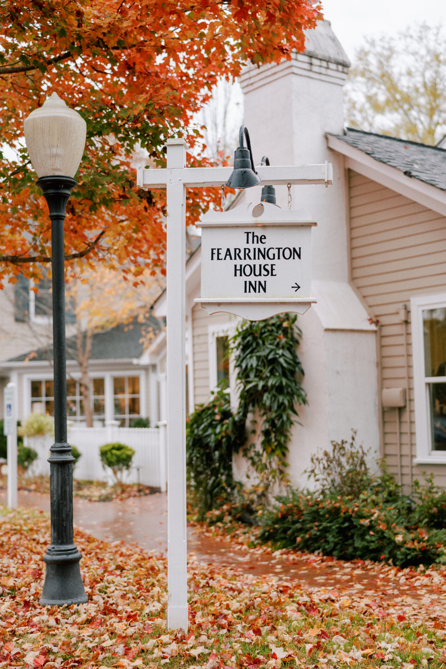 A quaint inn sign reading "The Fearrington House Inn" with an arrow pointing right, hangs from a white post beside a lamppost. Vibrant autumn leaves blanket the ground, and trees with orange foliage frame a charming, white cottage in the background. Fearrington Village