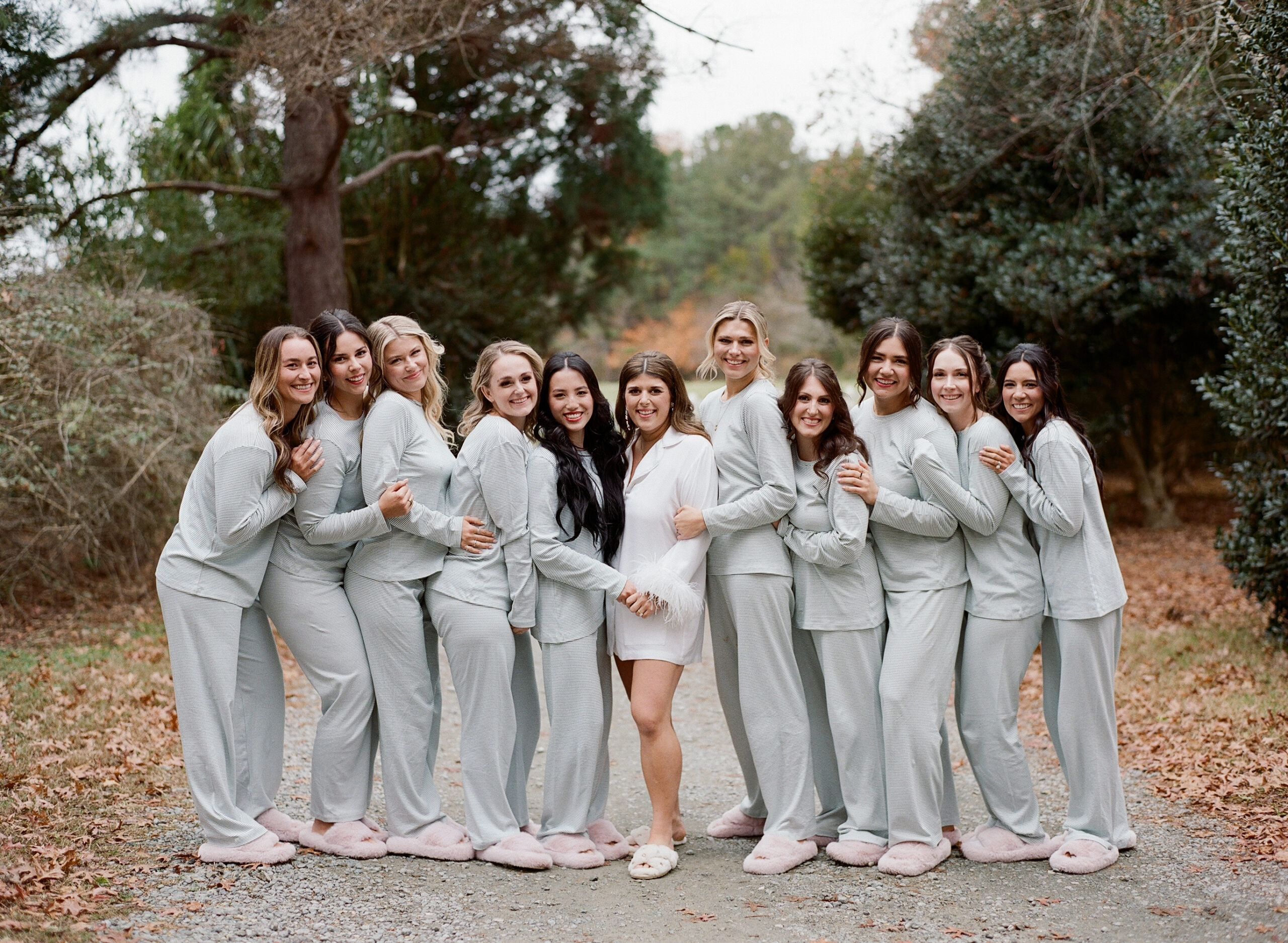 A group of eleven women are standing outdoors on a gravel path surrounded by trees, all wearing matching light-colored pajamas and slippers. The woman in the center wears a white pajama set. They are smiling and hugging each other, forming a line. Fearrington Village