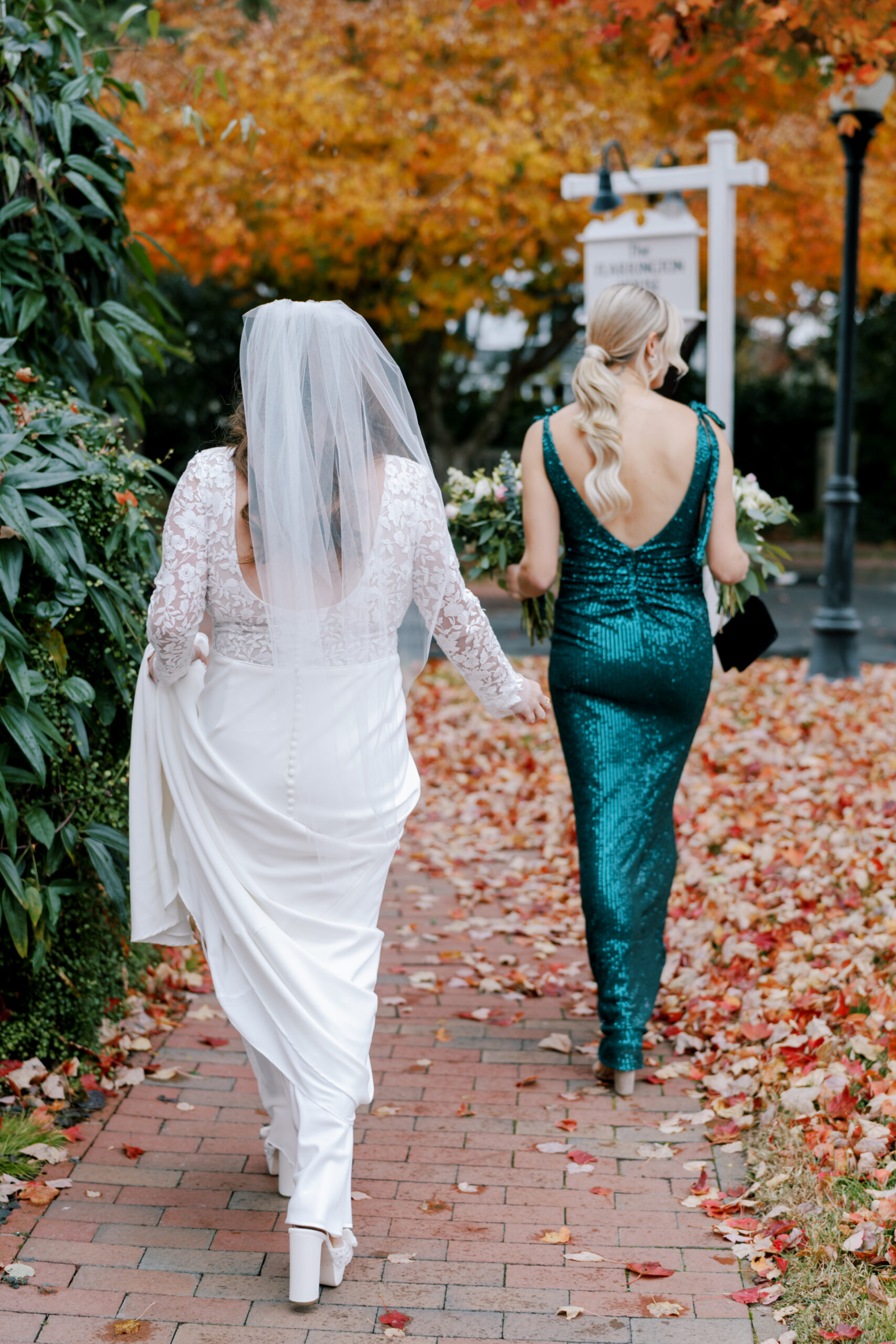 A bride in a white lace wedding dress and veil walks alongside a woman in a teal sequined gown. They are on a brick path strewn with autumn leaves, heading towards a signpost and a black street lamp, surrounded by trees with vibrant fall foliage. Fearrington Village