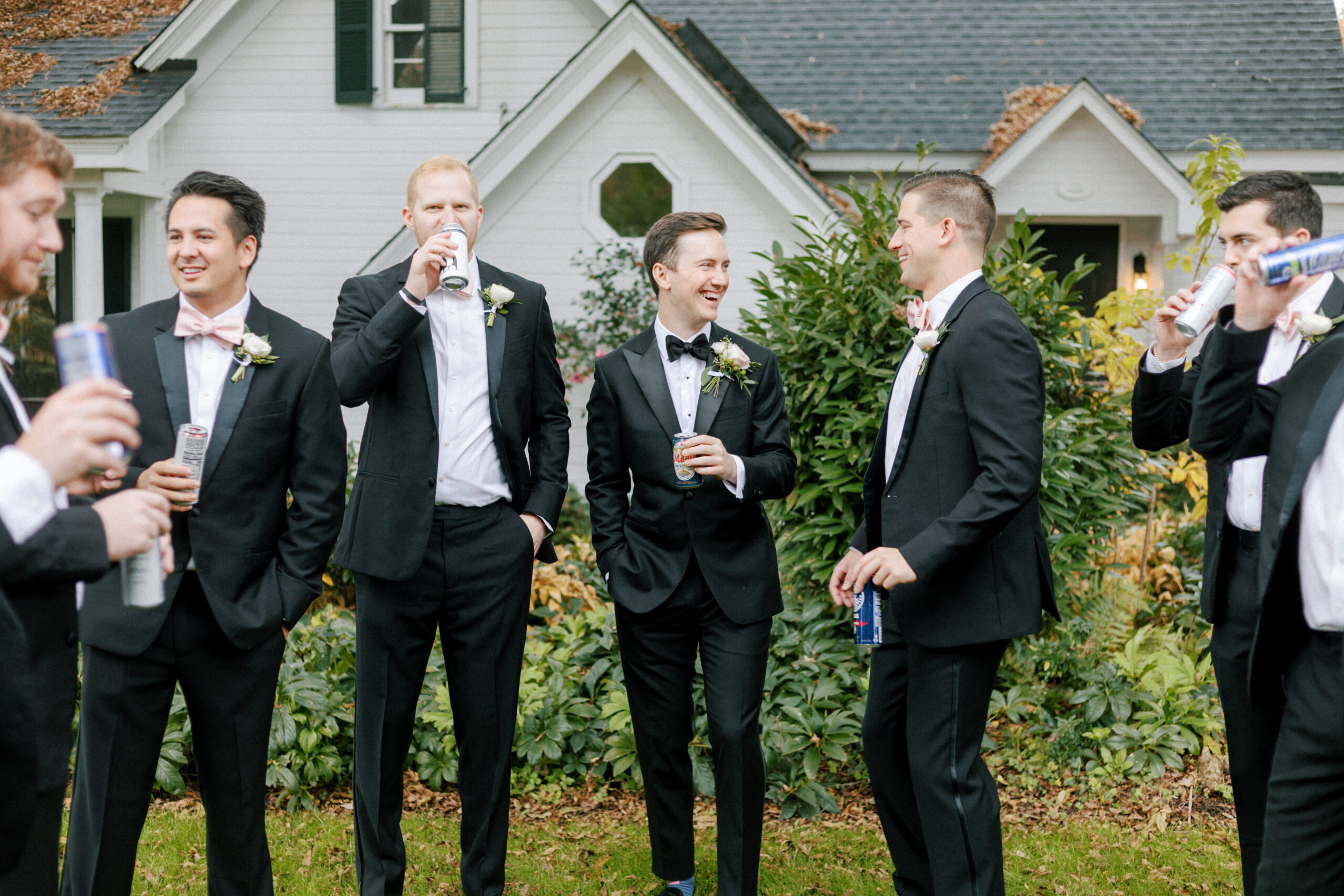 A group of men in black tuxedos with white shirts and black bow ties stand together outdoors, laughing and holding canned drinks. In the background, there's a white house with dark green shutters and a landscaped yard. The mood is cheerful and festive. Fearrington Village