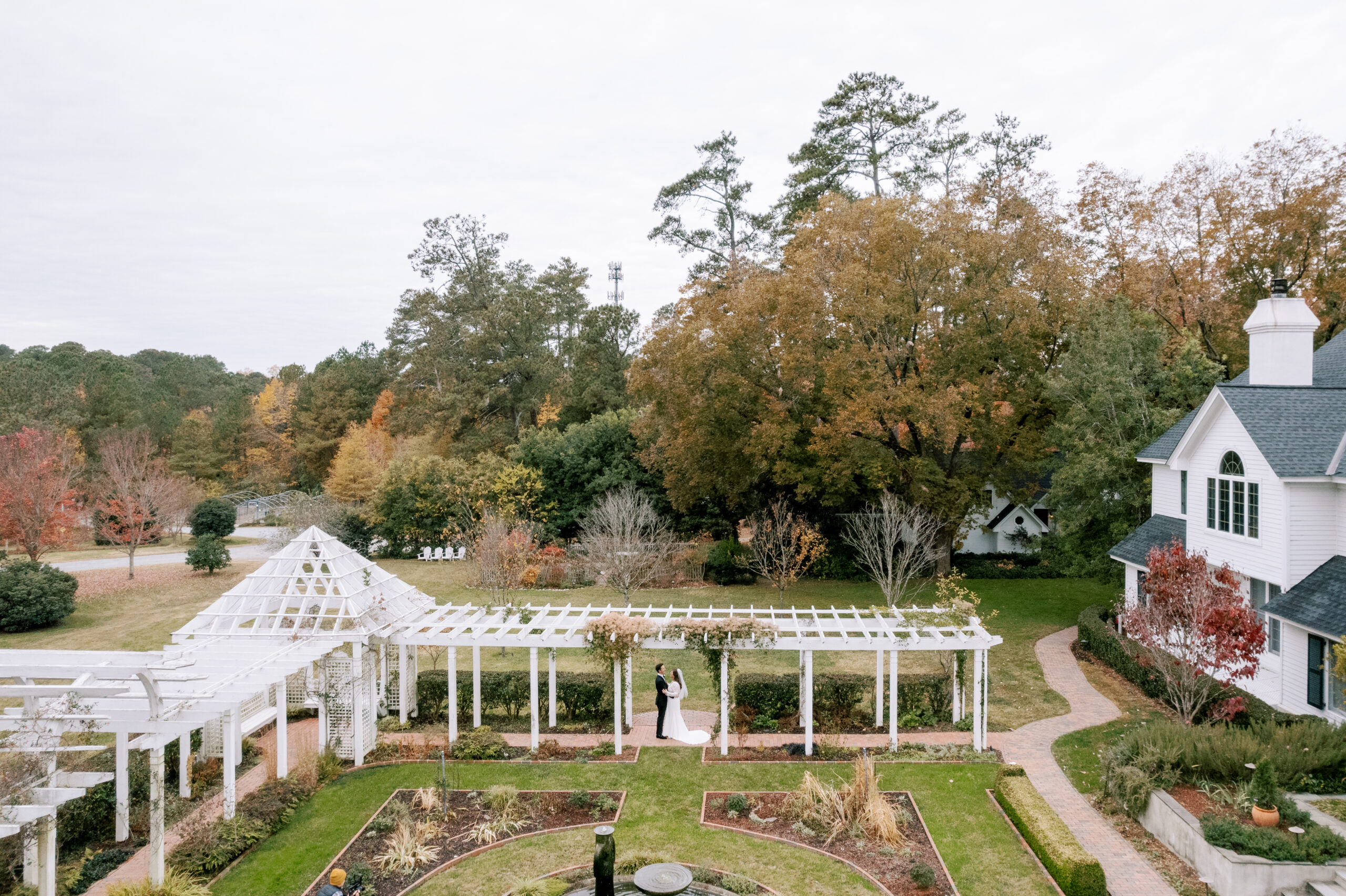 A couple stands under a white pergola in a garden with neatly arranged pathways and greenery. Nearby, a large house with a dark roof and white walls is surrounded by autumn trees, with various shades of orange, yellow, and green leaves. The sky is overcast. Fearrington Village