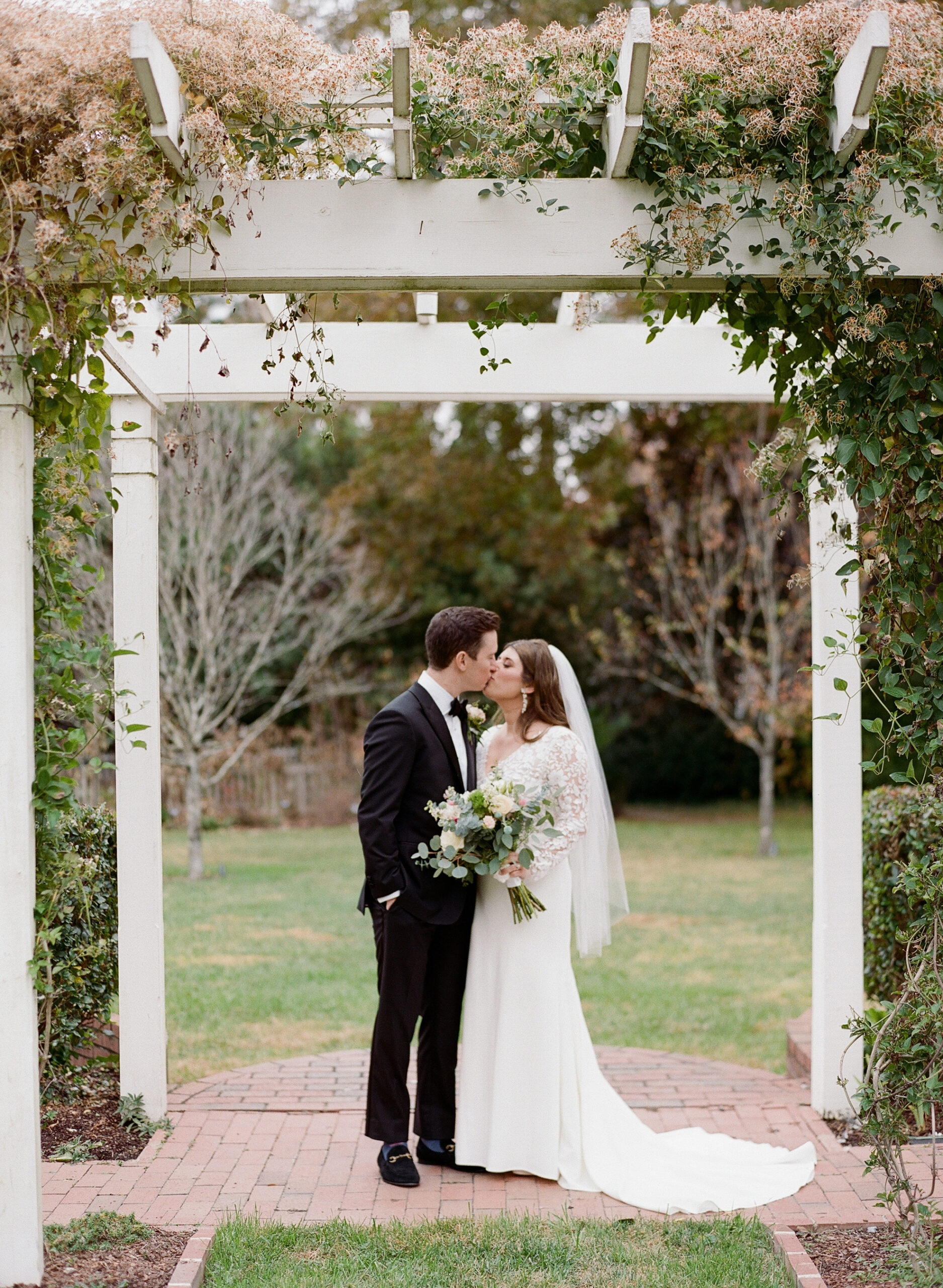 A newlywed couple shares a kiss beneath a white floral pergola in a garden setting. The bride is wearing a long white dress with lace sleeves and holding a bouquet of flowers, while the groom is dressed in a black suit and tie. Fearrington Village