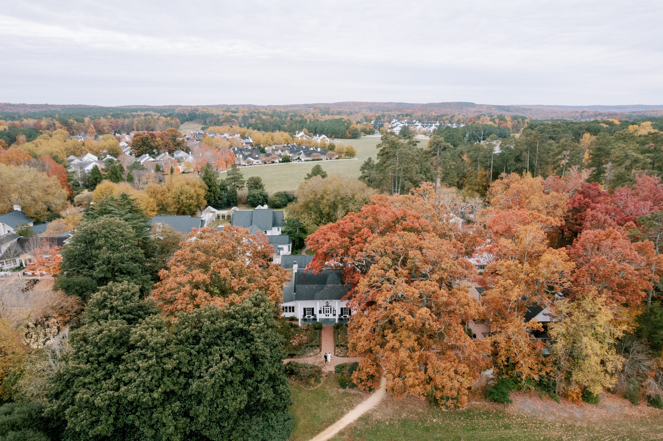 An aerial view of a suburban neighborhood in autumn. Houses are surrounded by trees with vibrant fall foliage in shades of red, orange, and yellow. A path runs through the area, with a mix of green spaces and residential streets visible in the background. Fearrington Village