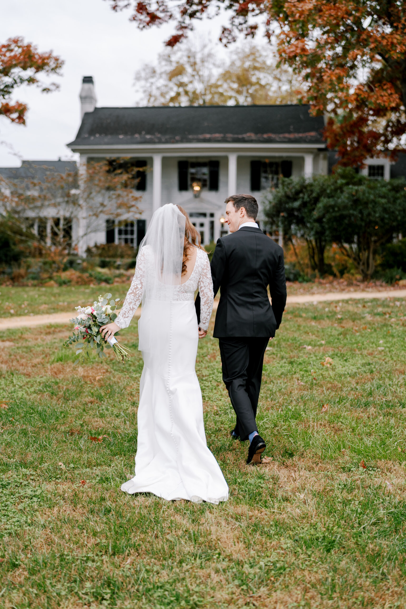 A bride and groom walk hand in hand towards a large, white house with dark shutters and a porch. The bride wears a long-sleeved white gown and veil, while the groom is in a black suit. They walk on a grassy lawn with autumn-colored trees surrounding the scene. Fearrington Village