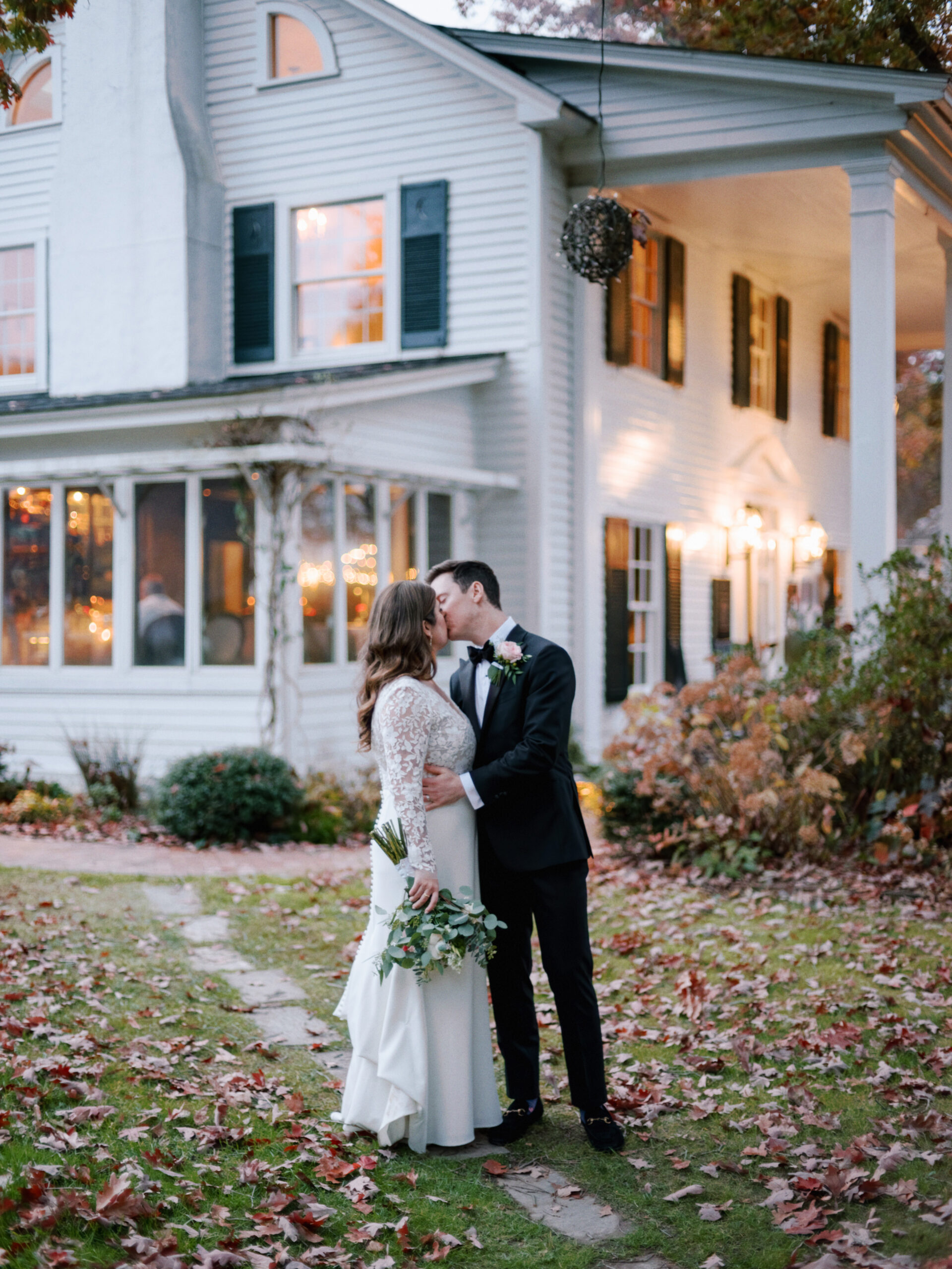 A couple in wedding attire share a kiss in front of a two-story white house with black shutters. The bride holds a bouquet and wears a lace dress, while the groom is in a black suit. The ground is scattered with fallen leaves, and the house's windows glow warmly. Fearrington Village