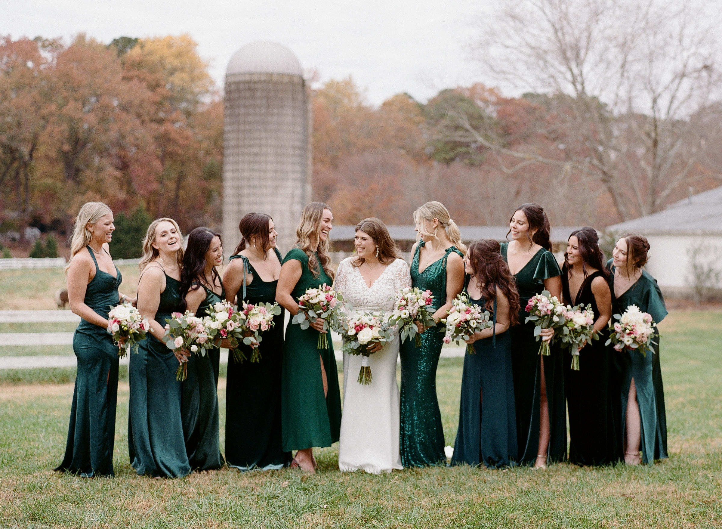 A bride in a white dress stands in a grassy area with ten bridesmaids in green dresses. They are holding bouquets of flowers and laughing, with a farm-like background featuring a silo, trees with autumn-colored leaves, and a white fence. Fearrington Village