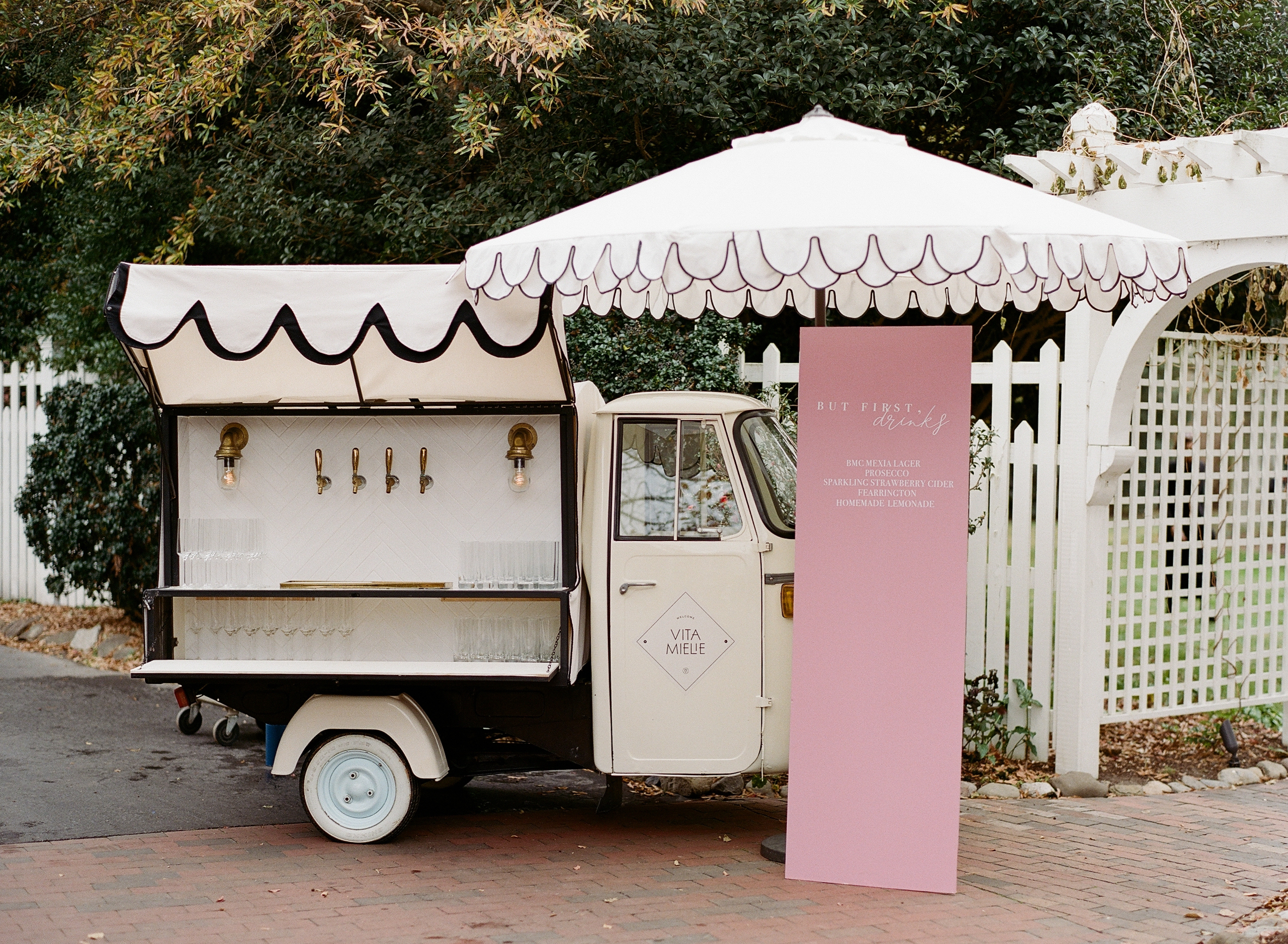 A retro-style mobile bar set up in a small three-wheeled vehicle with a pink and white canopy. An umbrella provides shade over the wooden countertop with four taps. A large pink sign beside it displays menu options. A white trellis and lush greenery are in the background. Fearrington Village