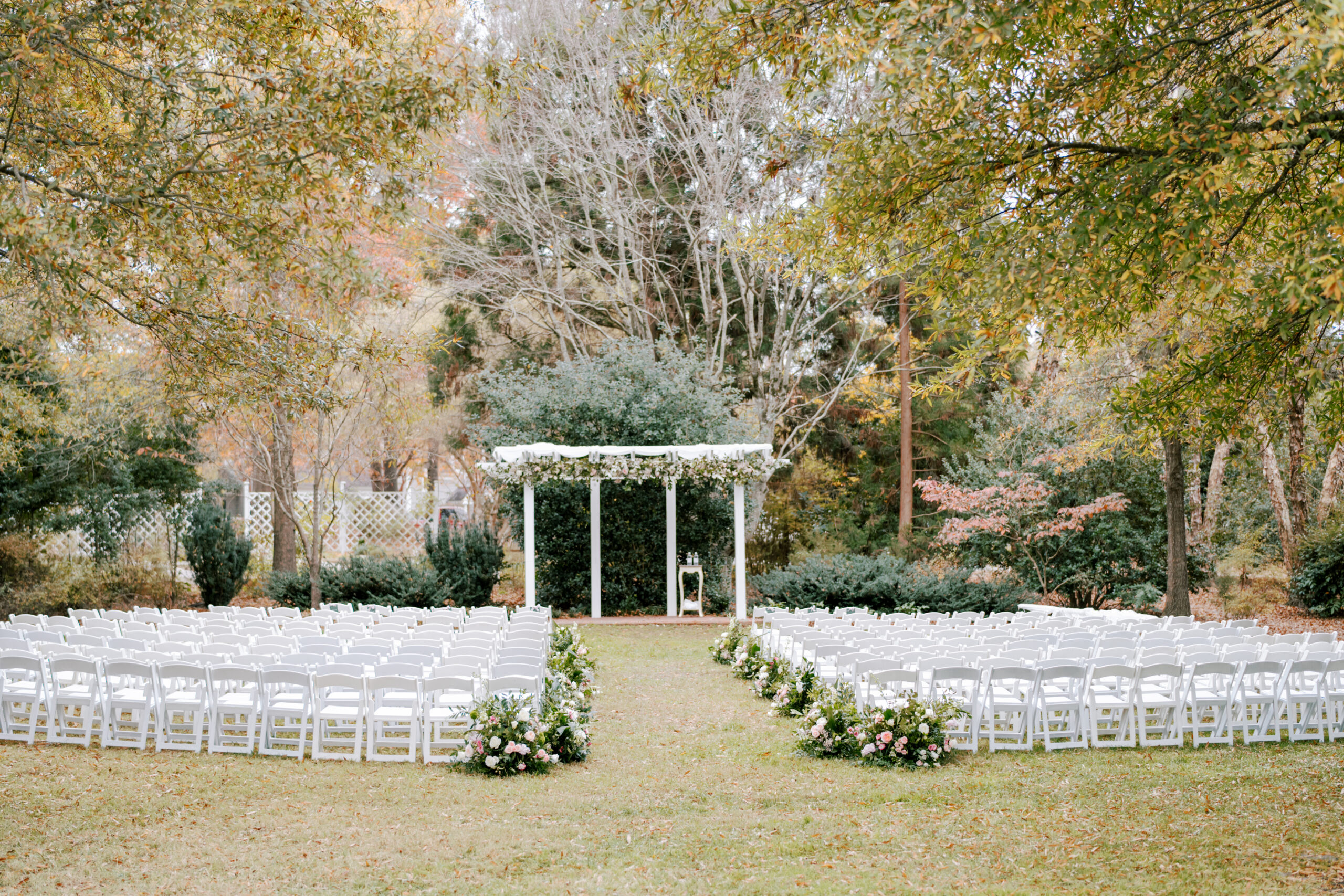 A picturesque outdoor wedding setup with rows of white chairs lined up facing a white arch adorned with greenery and flowers. The setting is surrounded by lush trees and garden greenery, creating a serene and romantic atmosphere. Fearrington Village